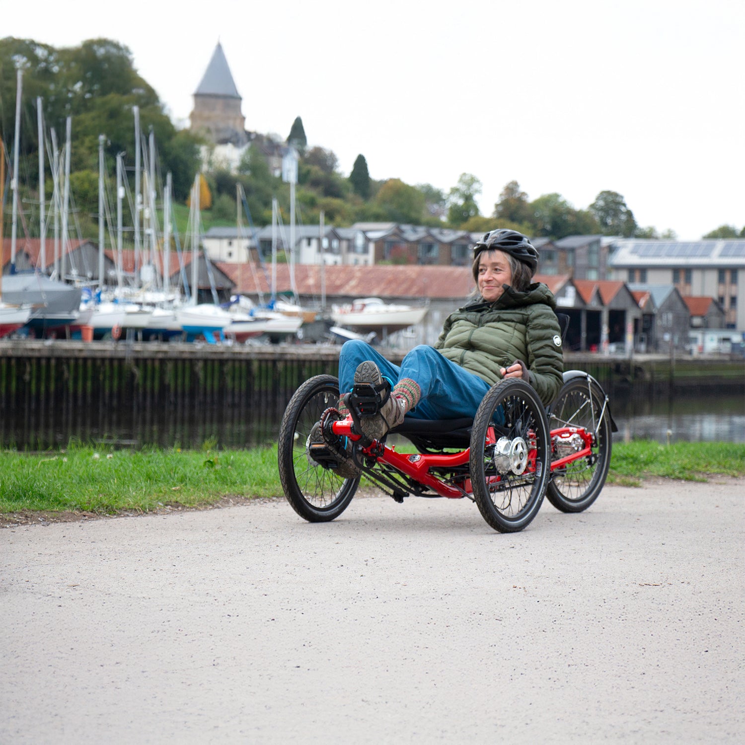 Person riding a red recumbent trike by a waterfront with boats and buildings in the background.