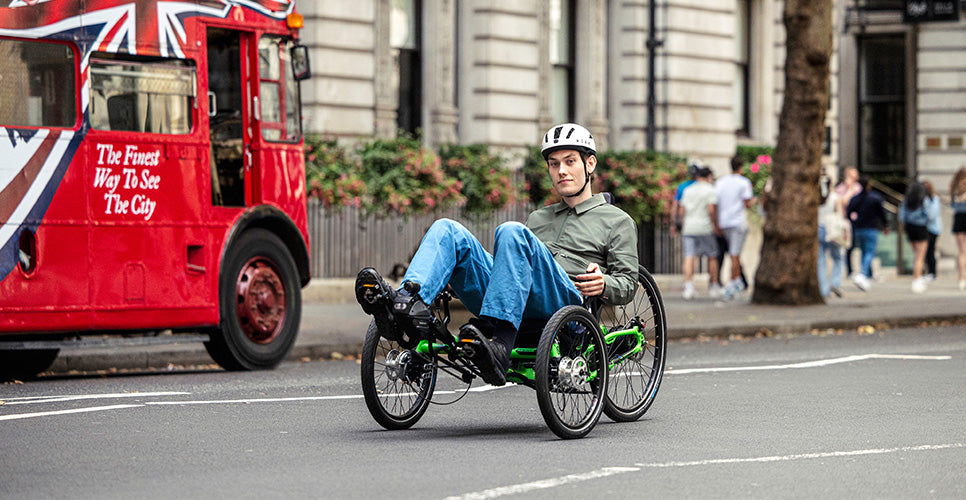 Person riding a recumbent trike on a city street with a red bus and pedestrians in the background.