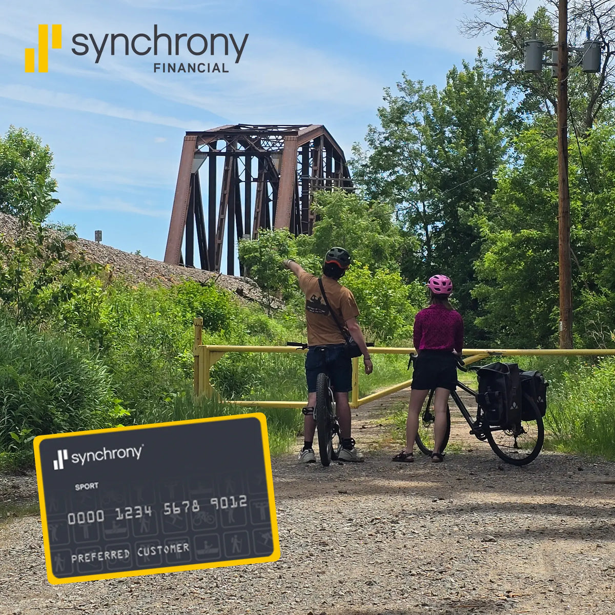 Two cyclists stop on a dirt path near a bridge, with one pointing toward the structure. A large black Synchrony credit card graphic is superimposed in the lower left corner.