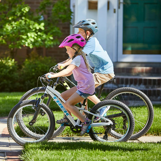 Two children riding bicycles on a sidewalk with a house in the background.