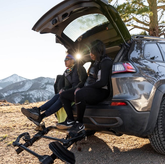 Kuat Transfer v2 Black 2 Bike Hitch Rack Fits 1.25 to 2 inch Receiver on a car tilted down with two people sitting on the tailgate.