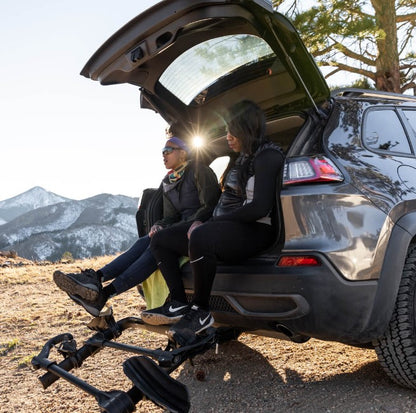 Kuat Transfer v2 Black 2 Bike Hitch Rack Fits 1.25 to 2 inch Receiver on a car tilted down with two people sitting on the tailgate.