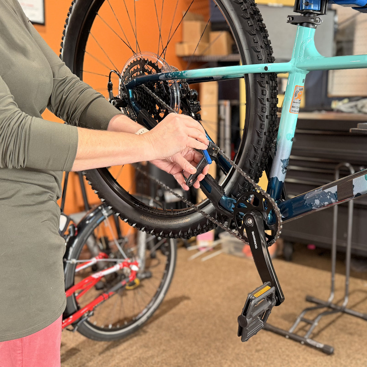 A close-up shot of a woman's hands show her working on the chain of a blue Trek mountain bike that's propped onto a bike stand.
