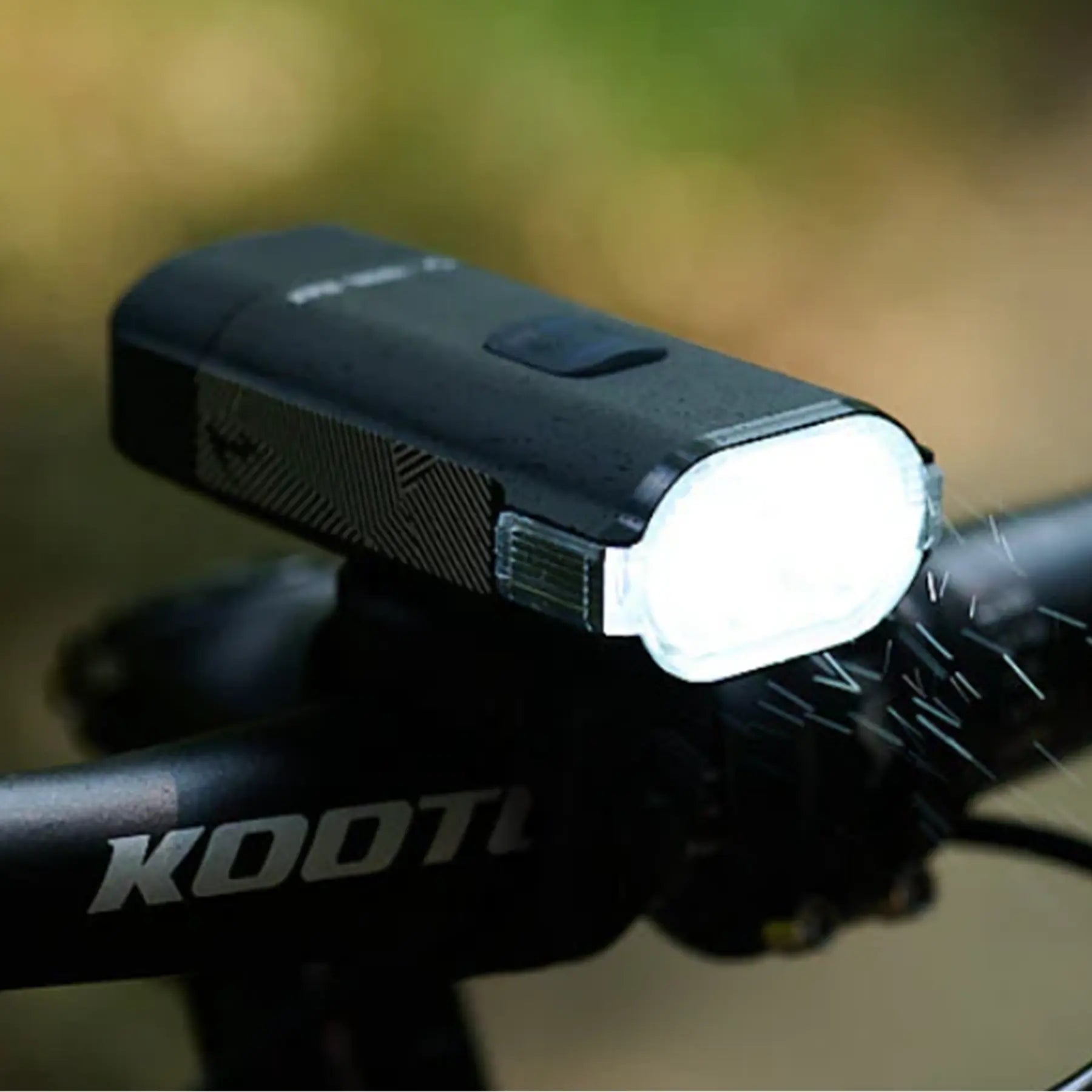 Image of the Moon Rigel Max 1500 Lumen Headlight in black turned on mounted on a bike handlebar against a blurry green background.