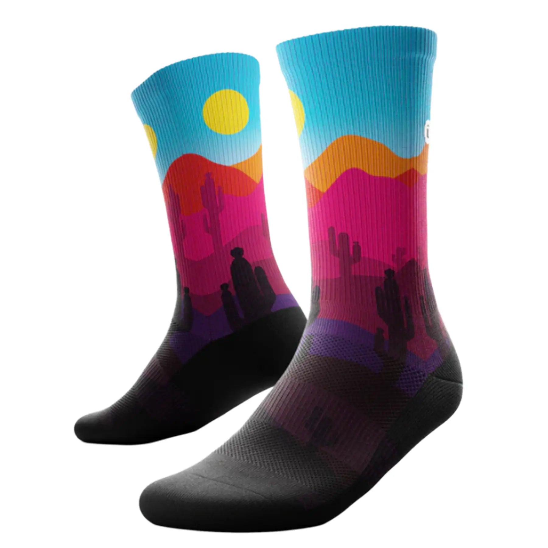 The Outway Dune Crew Socks feature a desert landscape with cactus in pinks, purples and orange, with a blue sky and a yellow sun, while the bottom of the sock is black.