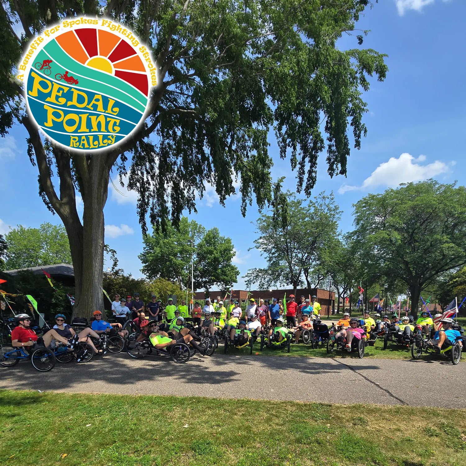 A large outdoor group photo at Pedal Point Bike Rally, featuring many cyclists in road bikes and hybrid bikes gathered under a large tree on a sunny day. A circular logo for 'Pedal Point Bike Rally' is overlaid in the top-left corner.