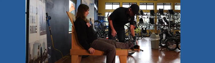 Person sitting on a wooden chair in a bike shop with another person working on sizing them.