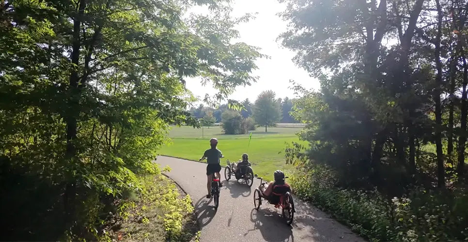 Two people riding trikes and one person riding a bike on a path through a park with trees and grass.