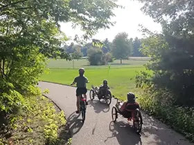 Two people riding rental tricycles and one person riding a rental bicycle on a paved path through a park with trees and grass.