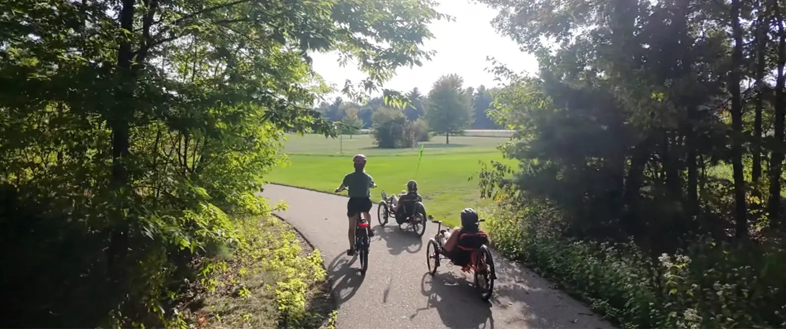 Two people riding rental tricycles and one person riding a rental bicycle on a paved path through a park with trees and grass.