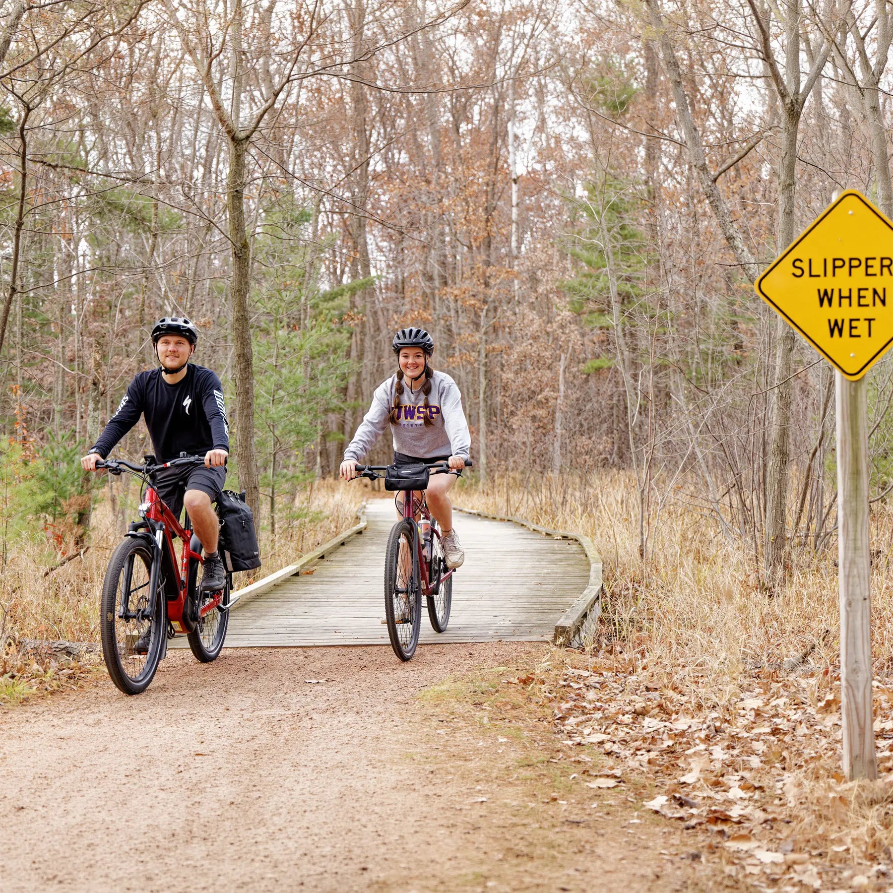 Two people riding bicycles on a wooden boardwalk trail in a forest with a 'Slippery When Wet' sign.
