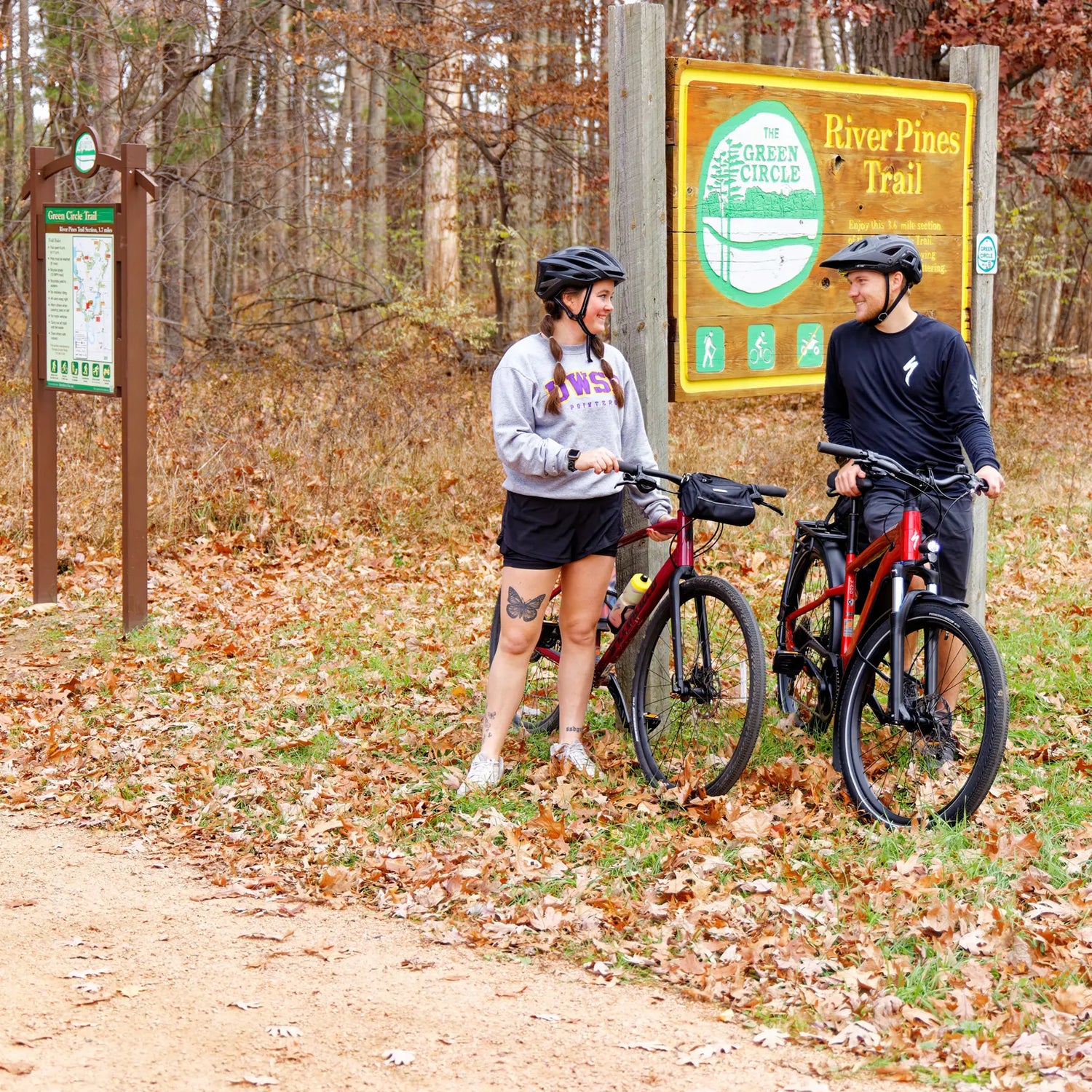 Two people with bicycles standing next to a Green Circle trail sign in a forested area.