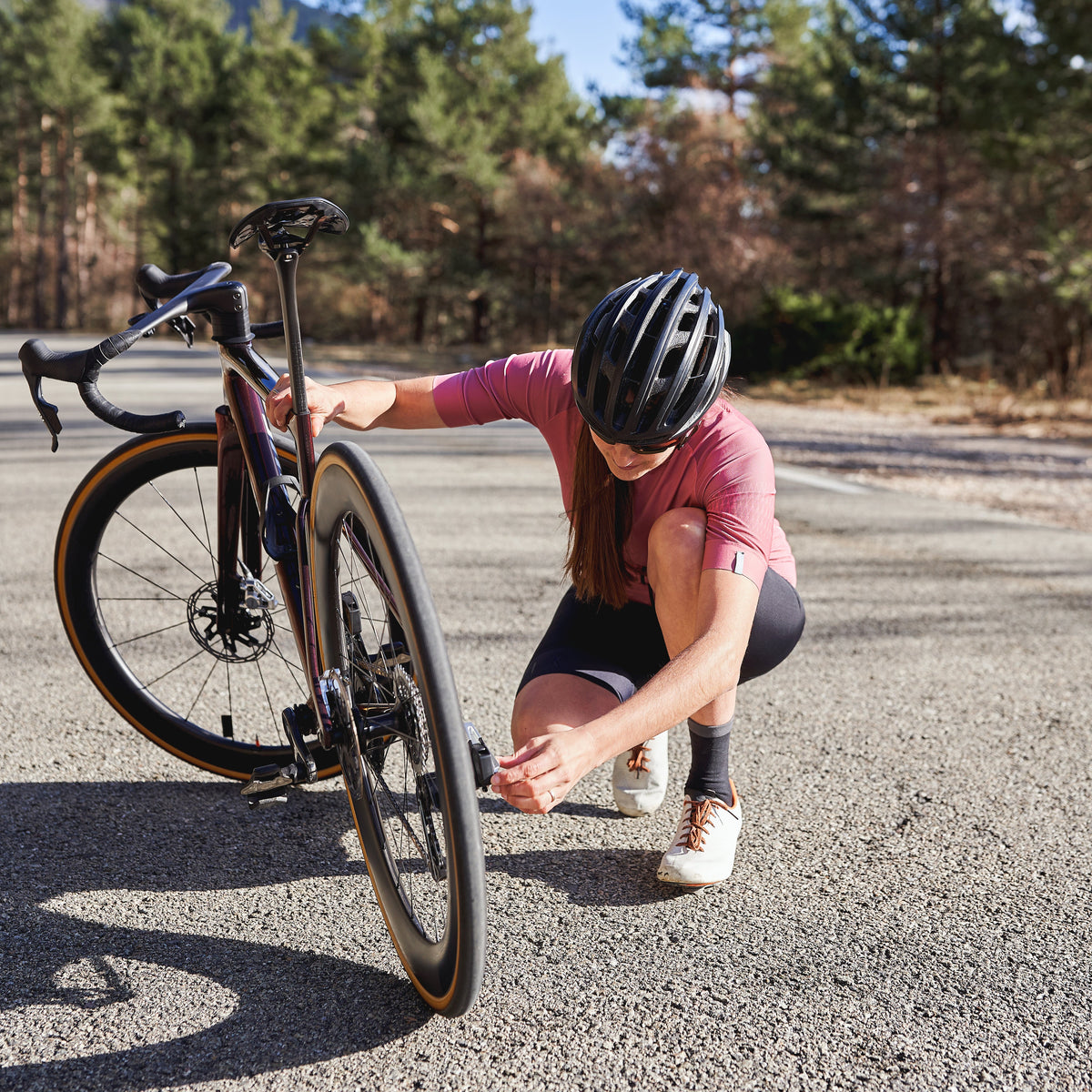 A cyclist in a pink jersey and black helmet crouches beside a road bike on an outdoor paved path, with trees and mountains in the background.