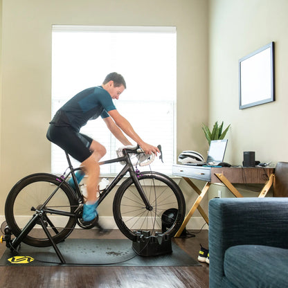 View of the Saris Indoor Bike Trainer Climbing Riser Block under a front bike wheel while a man rides his stationary trainer in his living room.