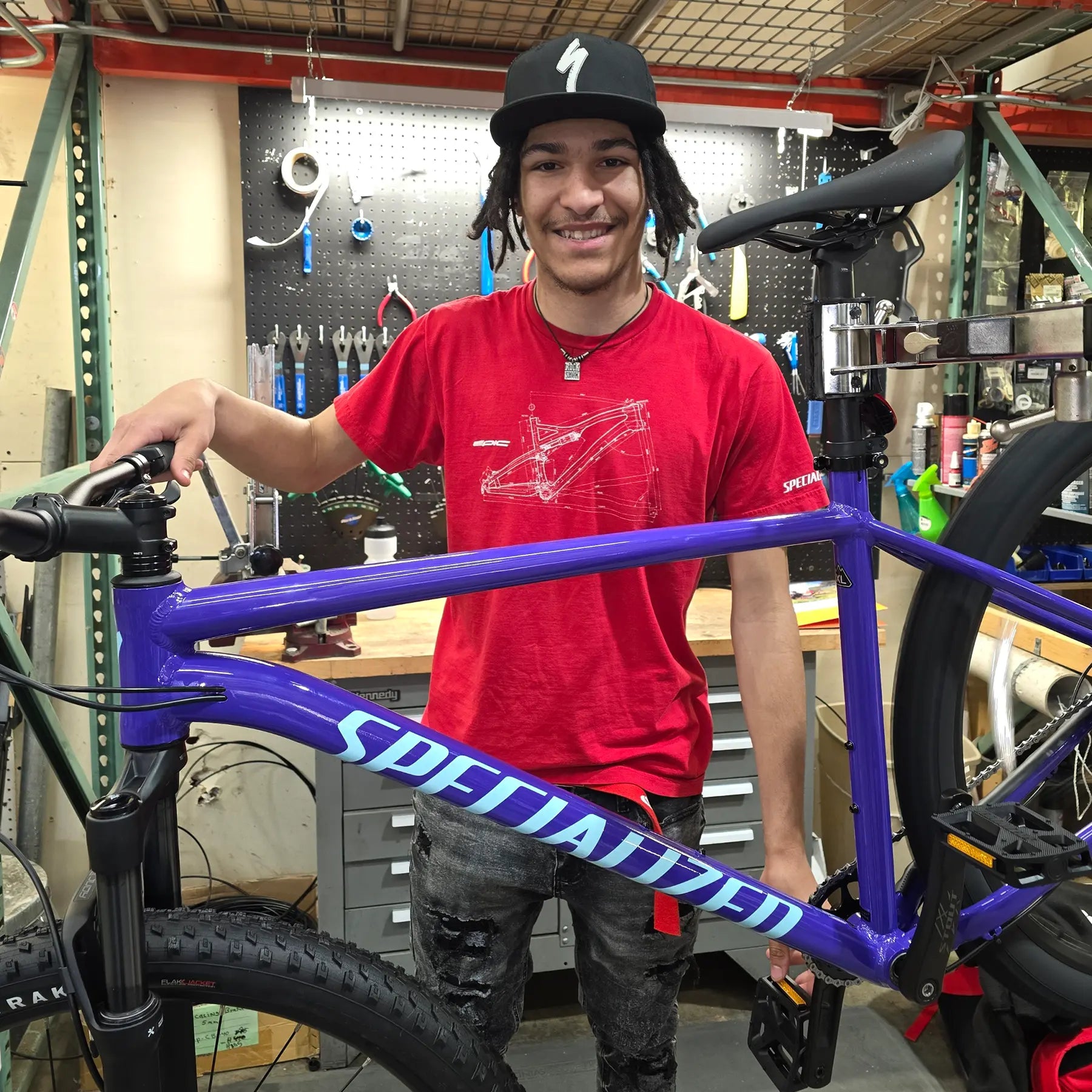 Person wearing a red shirt and black cap standing next to a purple bicycle frame in a workshop.
