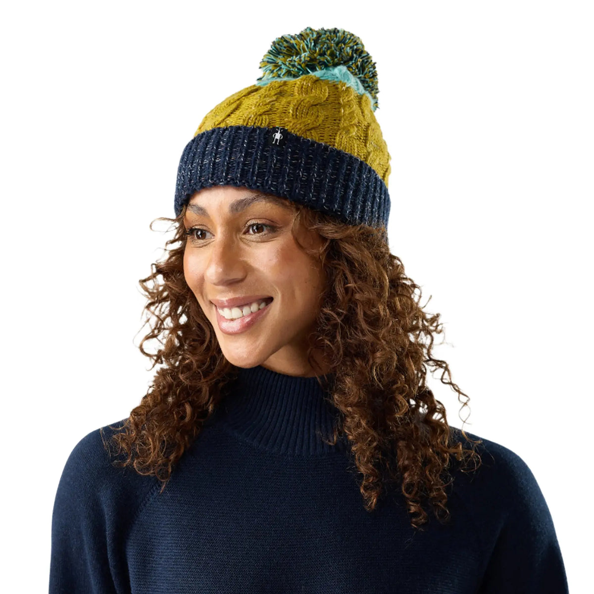 A smiling woman with curly brown hair wearing a knit Smartwool Isto Retro Beanieie in Bronze Olive Heather, paired with a navy turtleneck sweater, against a plain white background.