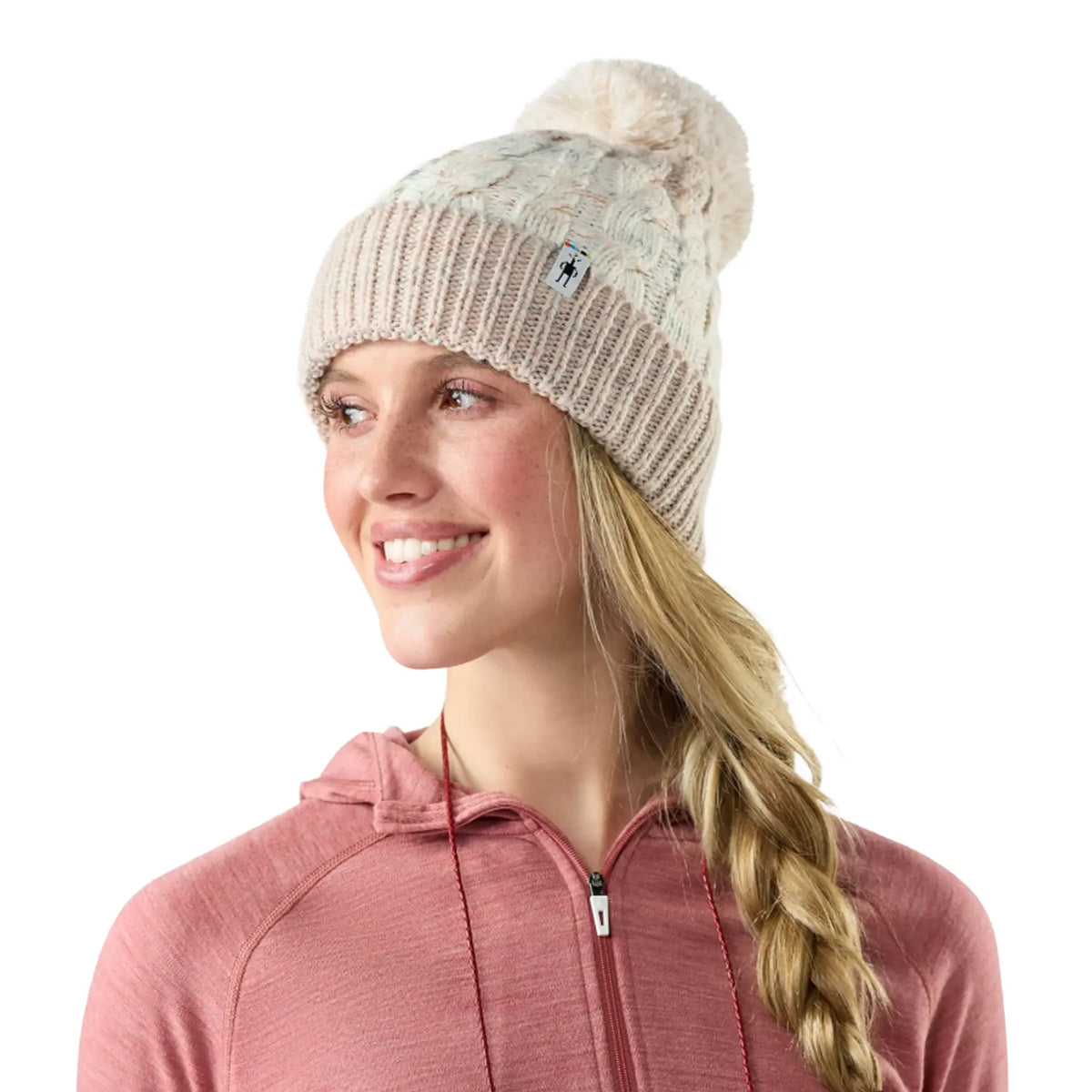 A woman wearing a Smartwool Isto Retro Beanie in Prosecco, a cream and beige cable-knit pom-pom beanie with a small logo patch, paired with a dusty rose zip-up hoodie, set against a plain white background. 