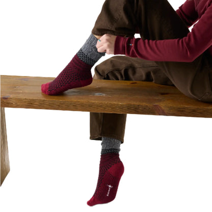 A woman sitting on a bench trying on a pair of Smartwool Womens Everyday Popcorn Cable Socks in Black and Tibetan Red, a grey and dark red sock with a black pattern against a white background.