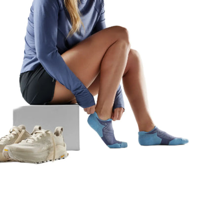 A woman in athletic wear sits on a white box, putting on a pair of Smartwool Women's Run Targeted Cushion Low Ankle Nightfall Blue Socks. A pair of off-white running shoes is placed on the floor beside her against a clean white background.