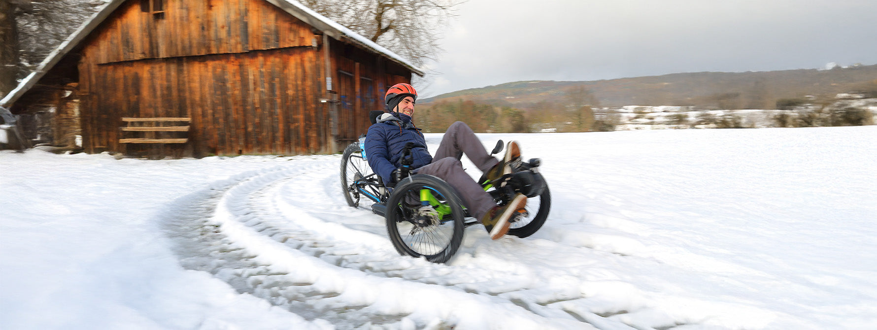 Person riding a recumbent trike on a snowy path with a wooden cabin in the background.