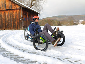 Person riding a recumbent trike on a snowy path with a wooden cabin in the background.