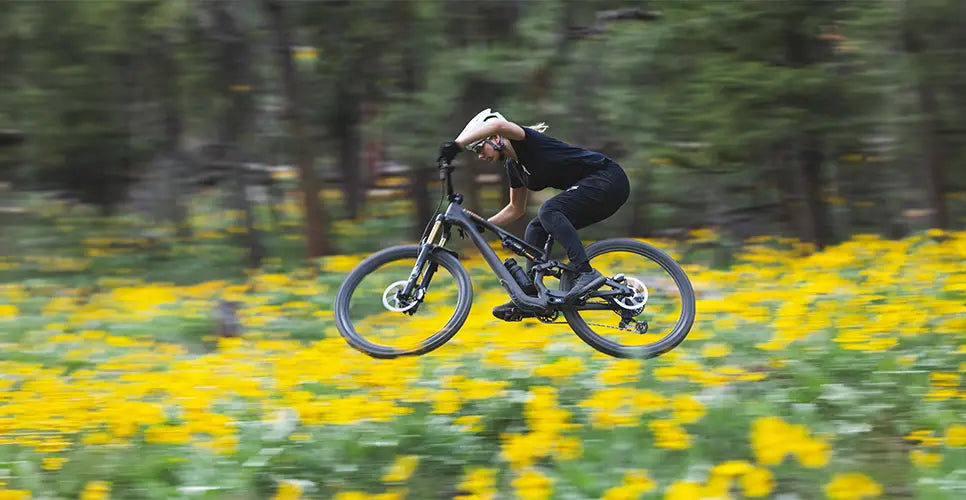 A woman rides a Specialized Mountain bike through a wooded area with yellow flowers in the foreground.
