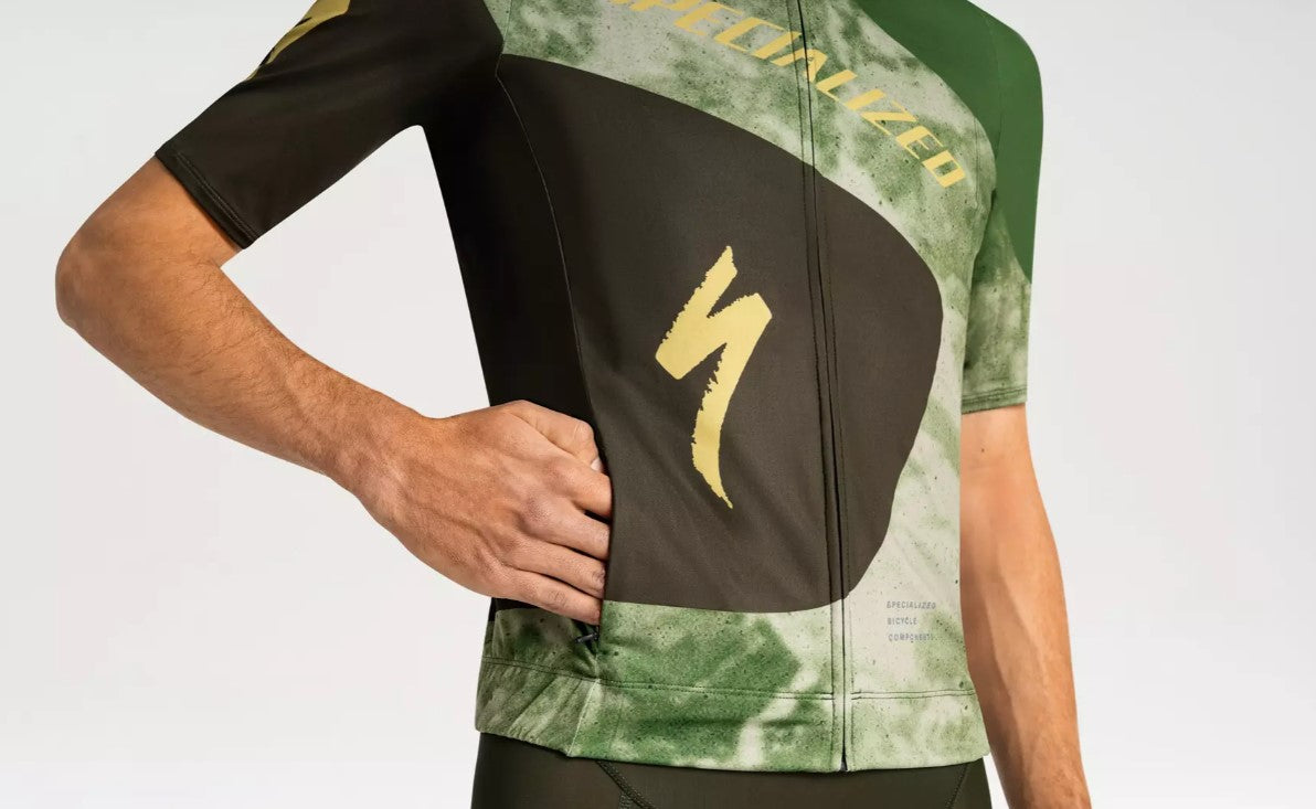 A close up view of a man putting his hand into a front right pocket of a green and black Specialized Foundation short sleeve cycling jersey on a white background.
