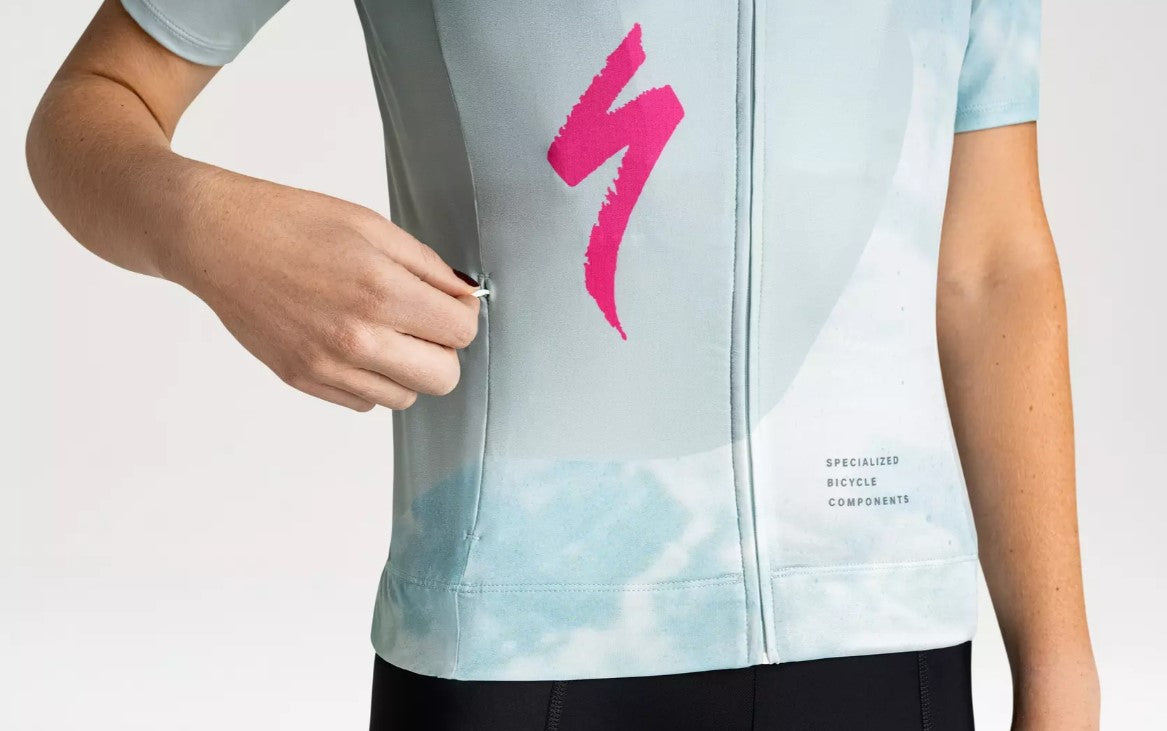 A close up view of a person unzipping a front pocket of a light blue Specialized Women's Foundation cycling jersey with pink Specialized logo on a white background.