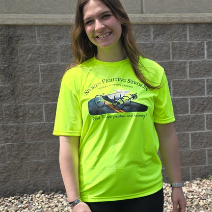 Closeup front view of the Spokes Fighting Strokes Cooling Performance Tee in Safety Yellow in an outdoor setting on a female model.