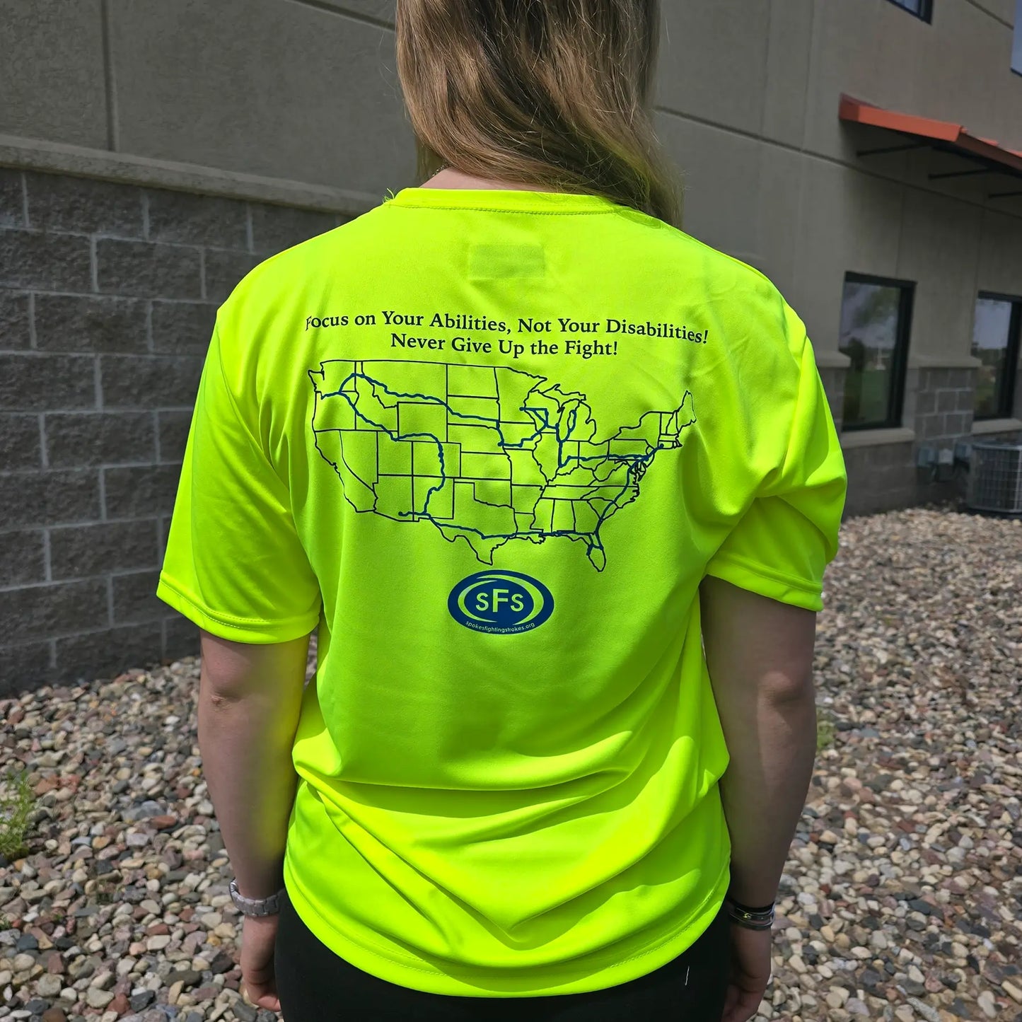 Closeup rear view of the Spokes Fighting Strokes Cooling Performance Tee in Safety Yellow in an outdoor setting on a female model.