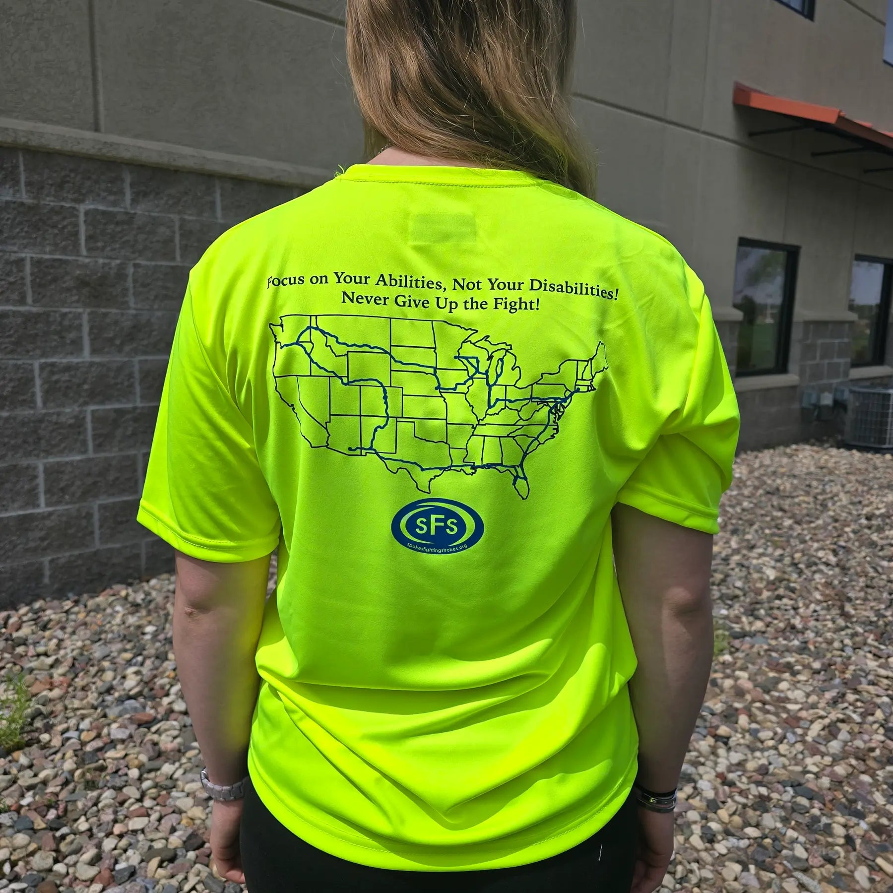 Closeup rear view of the Spokes Fighting Strokes Cooling Performance Tee in Safety Yellow in an outdoor setting on a female model.