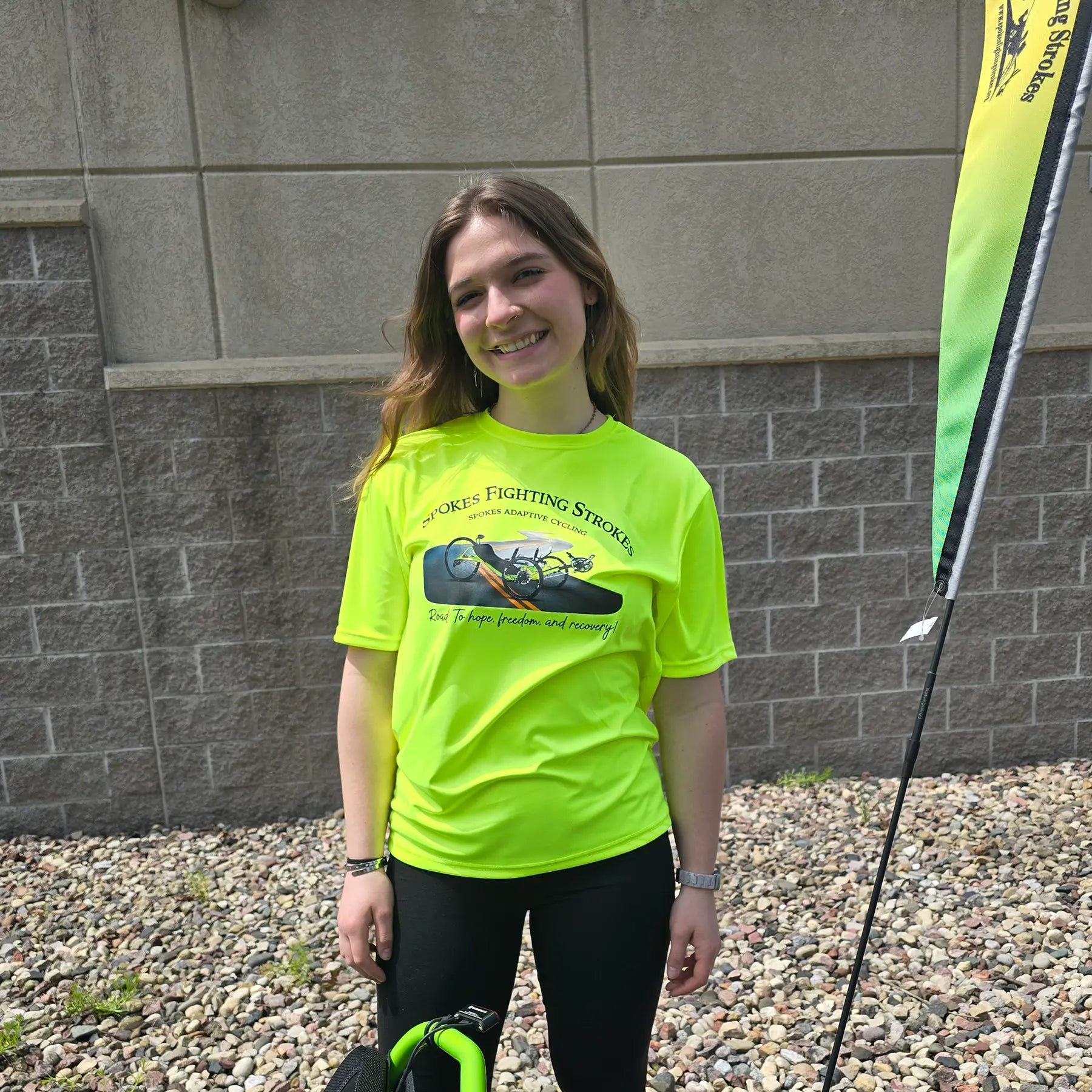 Front view of the Spokes Fighting Strokes Cooling Performance Tee in Safety Yellow in an outdoor setting on a female model.