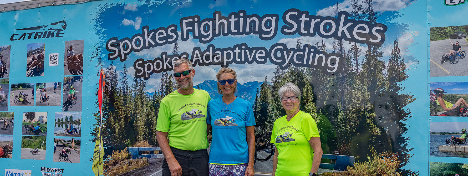 Three people standing in front of a promotional banner for Spokes Fighting Strokes and Spokes Adaptive Cycling.