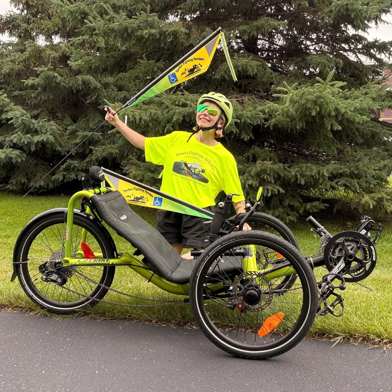 A person wearing a bright neon yellow-green Spokes Fighting Strokes t-shirt, matching helmet, and green sunglasses sits on a lime green and black recumbent trike on a paved path with evergreen trees in the background. The trike has a matching yellow-green and black Spokes Fighting Strokes flag attached to the handlebars.