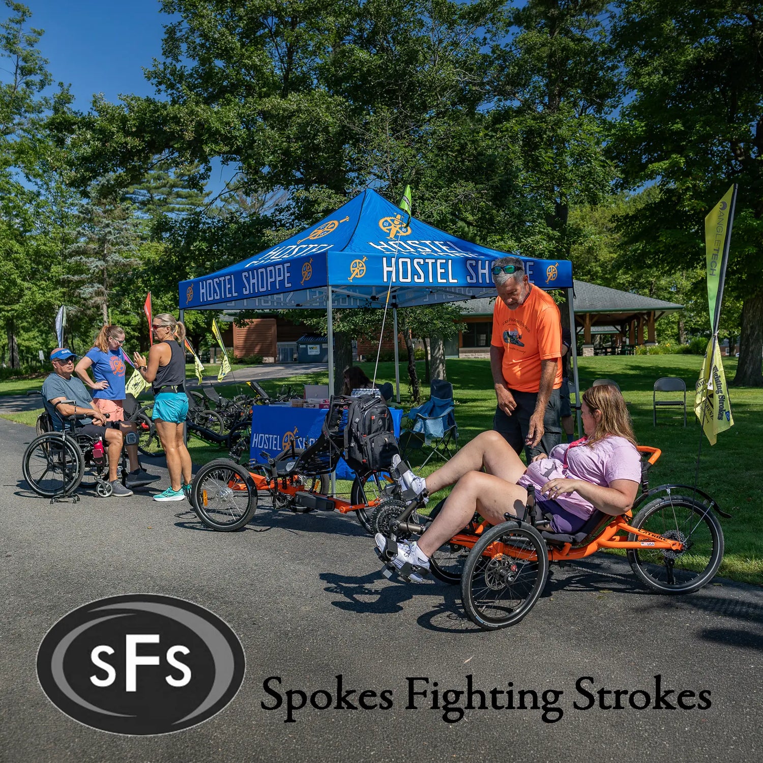 People using adaptive recumbent tricycles at an outdoor event with a 'Hostel Shoppe' tent in the background.