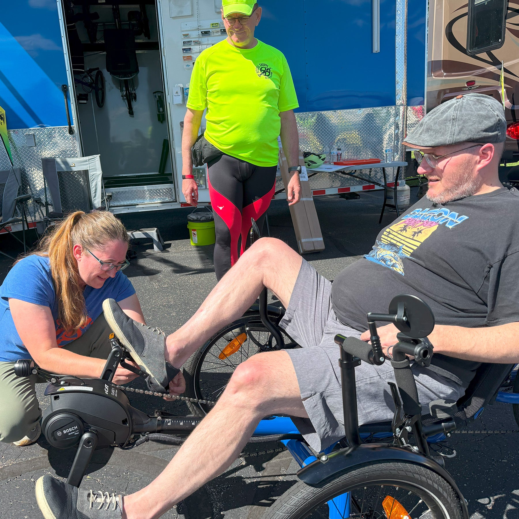 Person using a recumbent trike with another person assisting them, near a trailer.