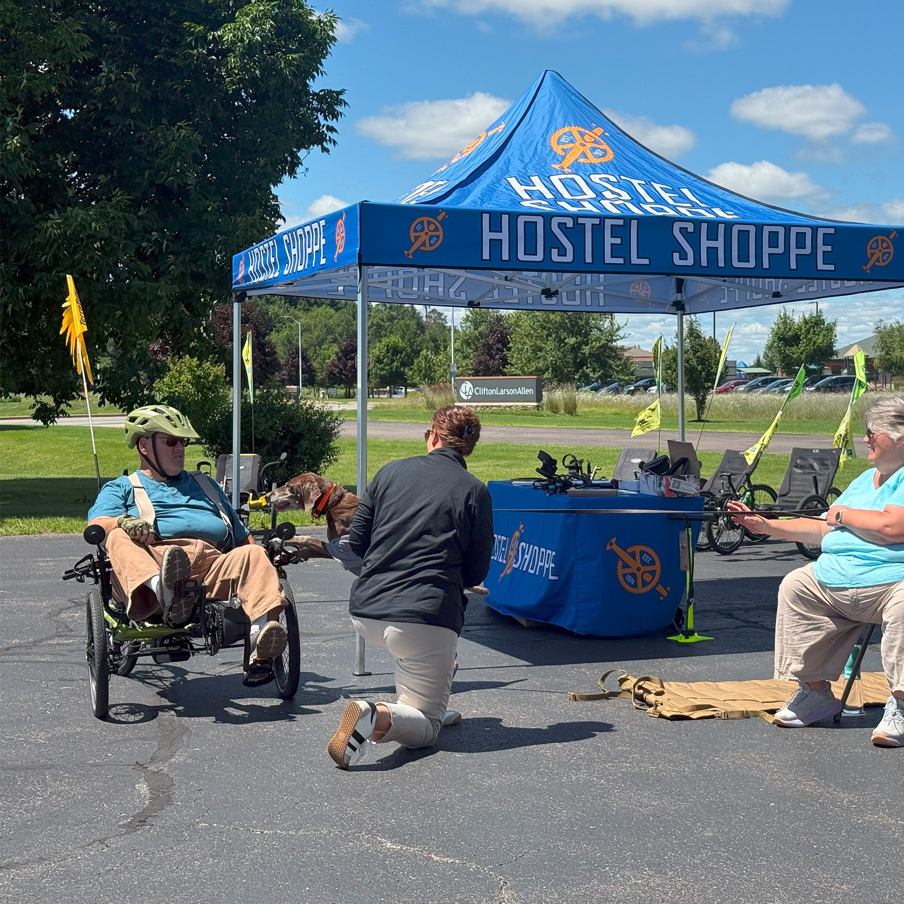 Person in a recumbent trike interacting with another person at a Hostel Shoppe event under a blue tent.