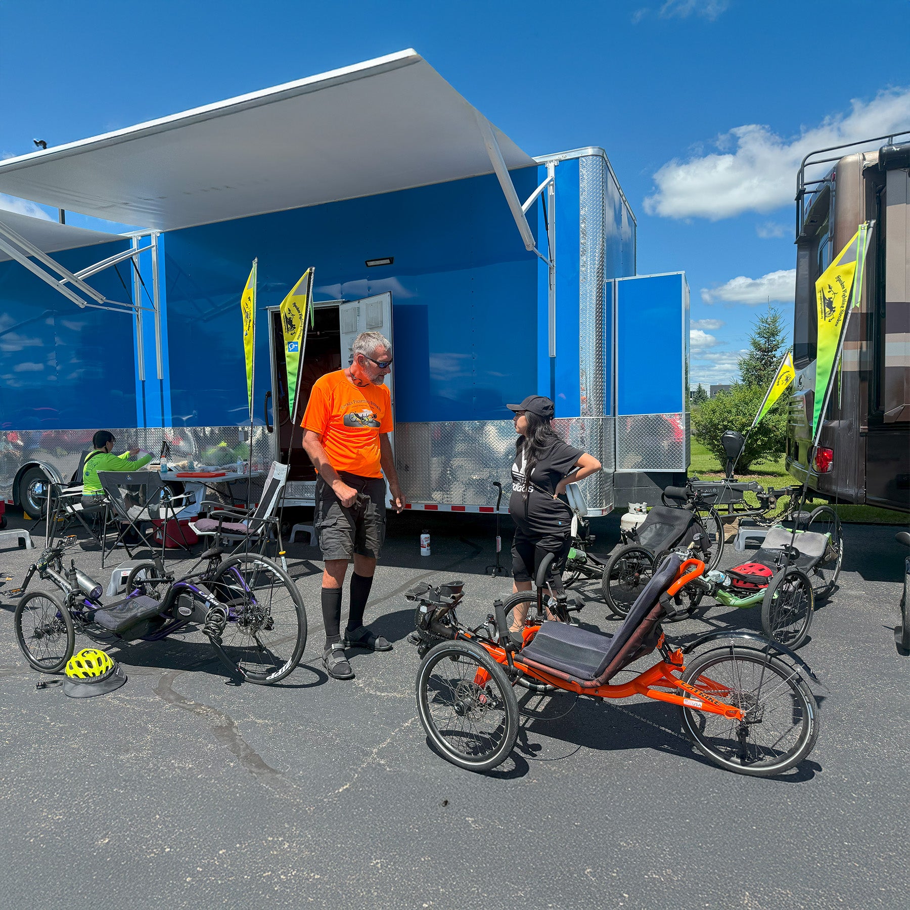 Two people standing next to recumbent trikes in front of a blue trailer on a clear day.