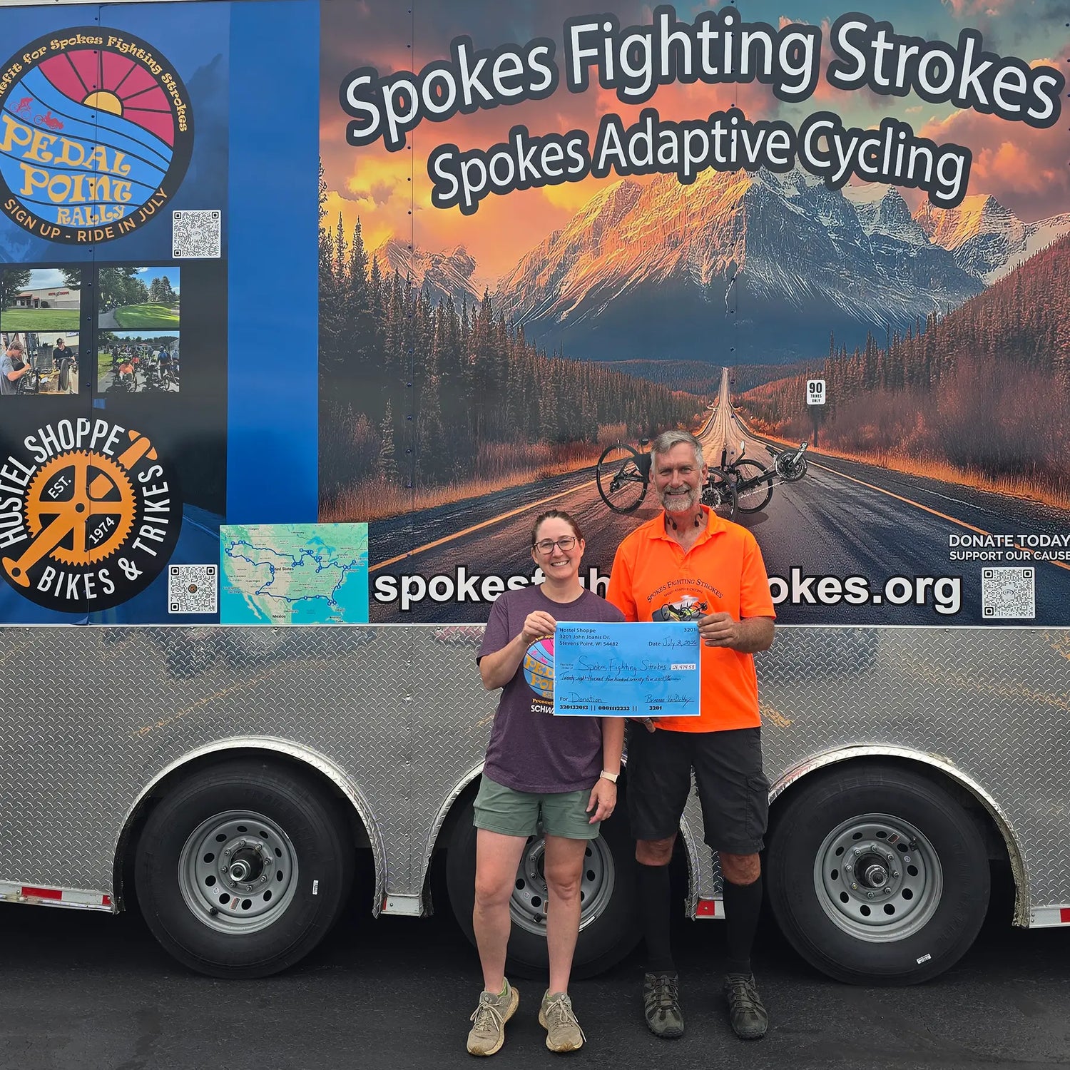 Hostel Shoppe owner, Breanna, and Dan Zimmerman from Spokes Fighting Strokes stand in front of the Spokes Fighting Strokes Trailer, with a fundraising check.