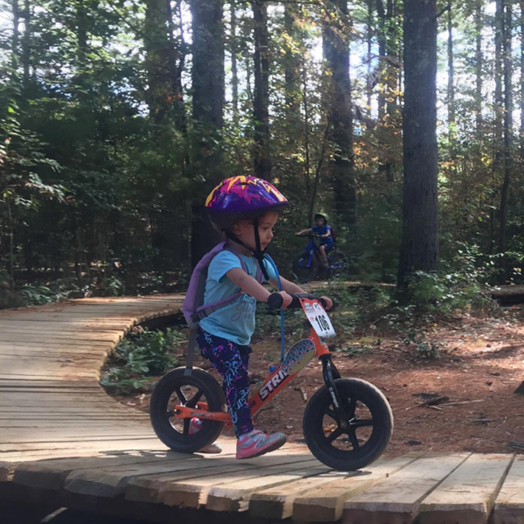 A toddler scoots along a wooden boardwalk on a Strider balance bike.