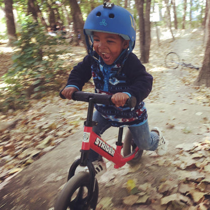 A toddler scoots down a dirt trail on a red Strider balance bike.