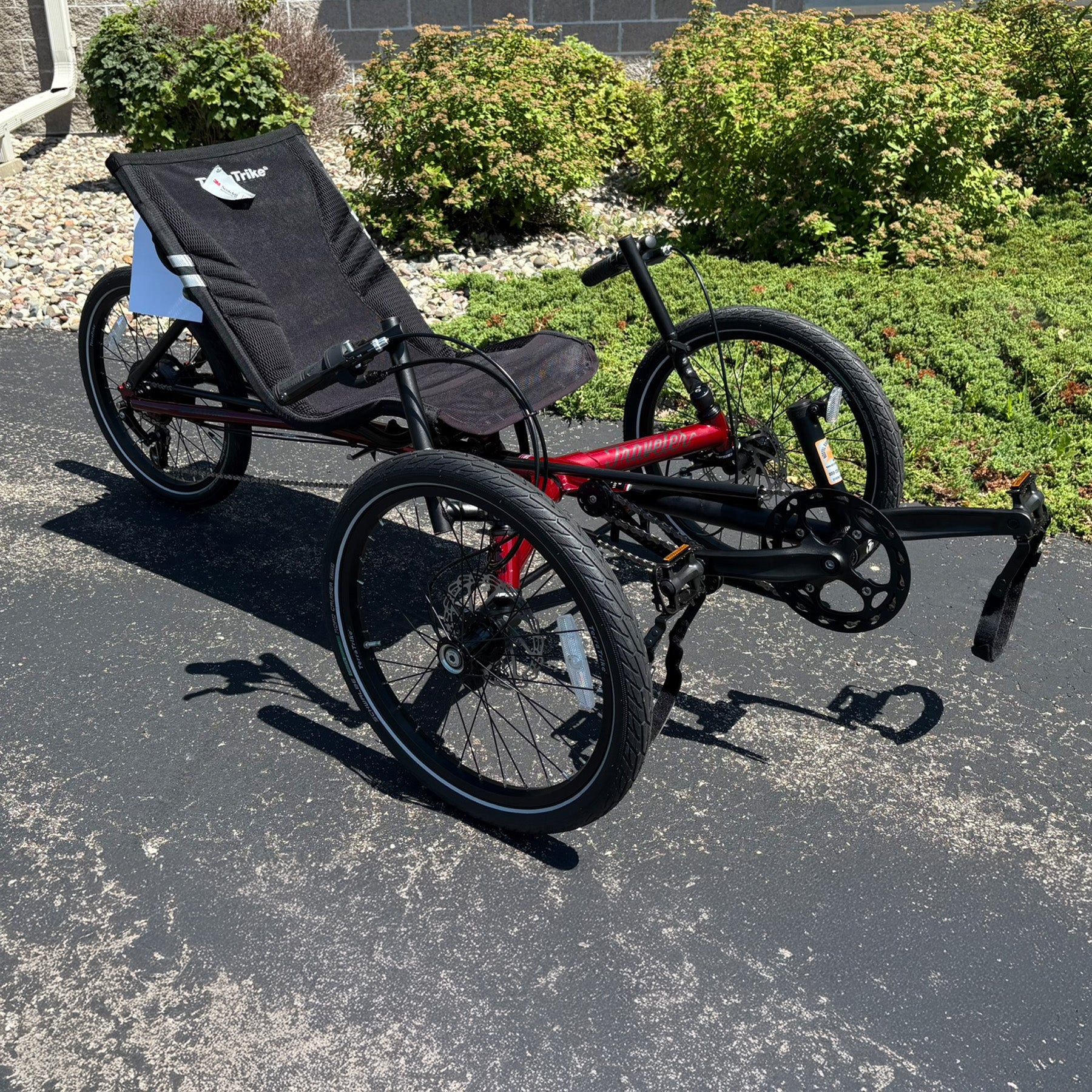 Angled view of a three-wheeled recumbent trike on a paved surface with greenery in the background.