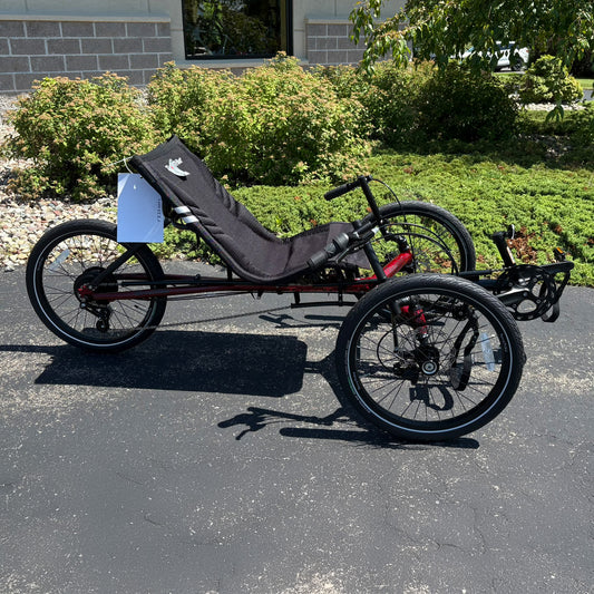Three-wheeled recumbent trike on a paved surface with greenery in the background