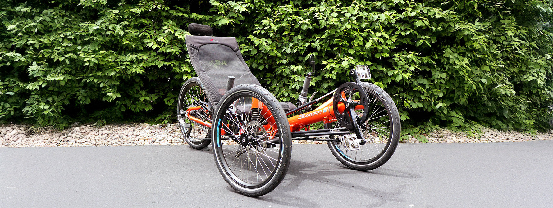 Three-wheeled orange recumbent trike on a paved path with greenery in the background.