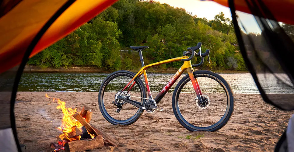 Image of a yellow, red and black Trek Road Bike framed by a tent opening next to a fire on a beach.