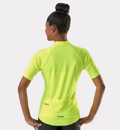 A woman shown from the back wearing a bright radioactive yellow Trek Solstice short-sleeve cycling jersey with a large rear pocket. The brand name TREK is printed in black on the pocket.
