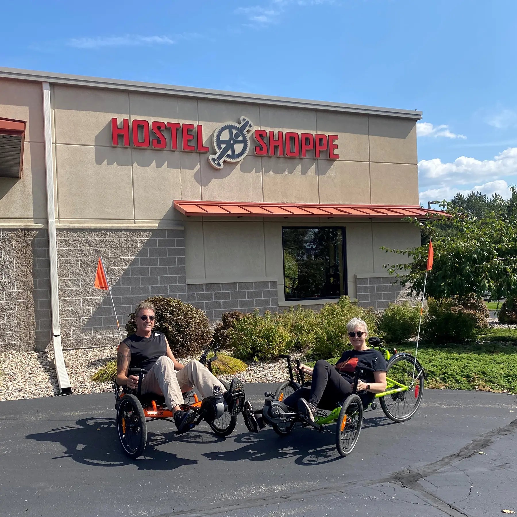 Two people on recumbent tricycles in front of the 'Hostel Shoppe' sign.