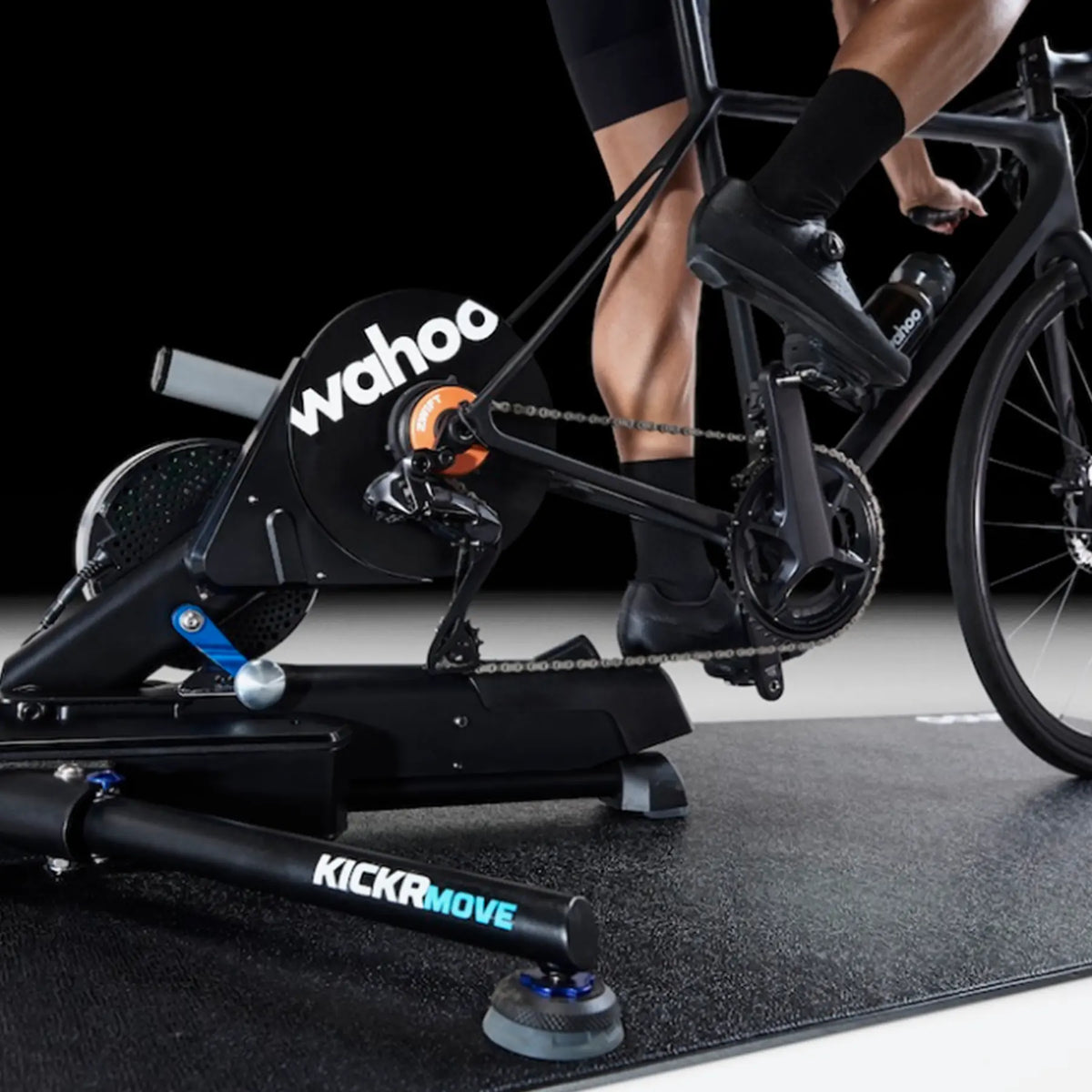 A Wahoo KICKR Bike Trainer Floor Mat underneath a Wahoo KickR Ride indoor cycling trainer attached to a road bike, with a rider's legs and feet visible in black cycling shoes and socks against a dark studio background.