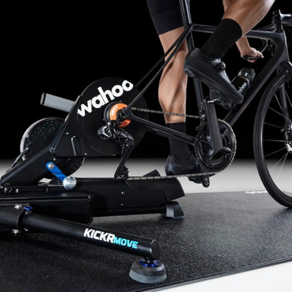 A Wahoo KICKR Bike Trainer Floor Mat underneath a Wahoo KickR Ride indoor cycling trainer attached to a road bike, with a rider's legs and feet visible in black cycling shoes and socks against a dark studio background.