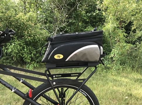 Lifestyle image of TerraTrike Deluxe Trunk Pack installed on a recumbent trike rear rack in the foreground and green grass and woody vegetation in the background. Bag is mainly black with silver and white reflective accents and a yellow and black TerraTrike oval logo on the side.